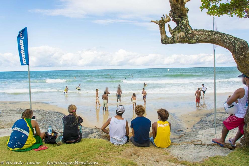 Imagem imagem de pessoa sentados na paria vendo surfistas no mar.